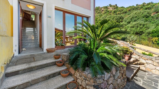 Un bâtiment à deux étages, doté d’un escalier en pierre menant à une porte en bois, entouré de plantes en pot et offrant une vue sur une colline.