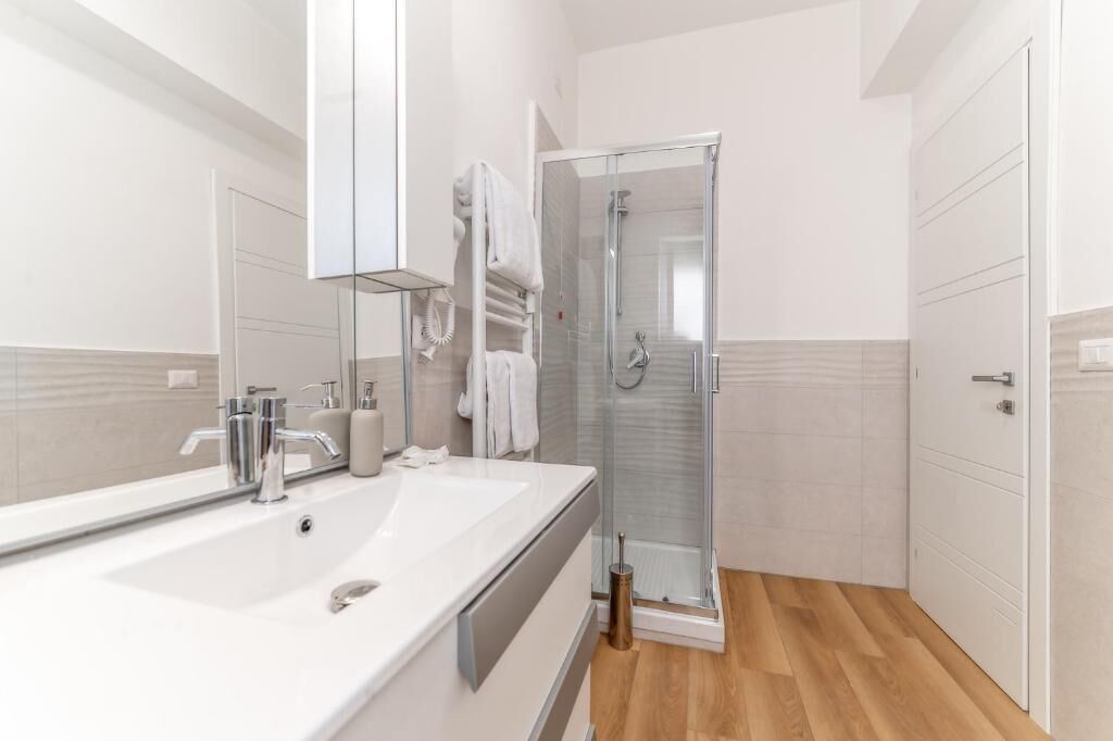 A modern bathroom with a large mirror, a white sink, a glass shower enclosure, and a wooden floor.