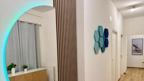 A hallway with a wooden floor, a radiator, a white cabinet with plants, a circular mirror with a light, and a wall with a geometric art piece.