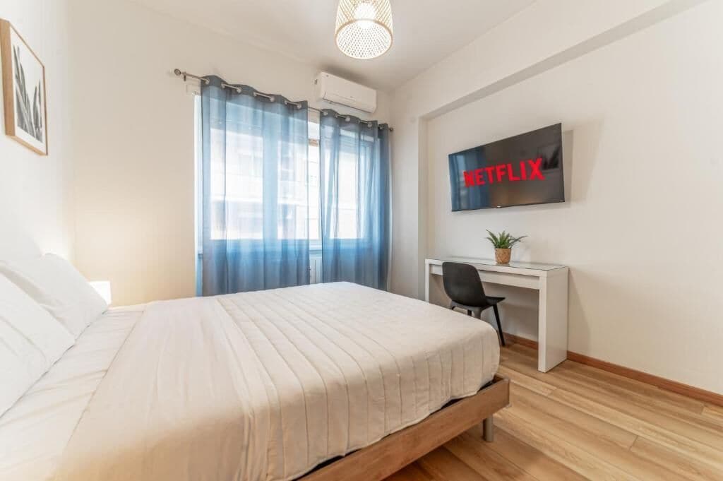 A bedroom with a bed, a desk, a chair, a TV, and a window with curtains.