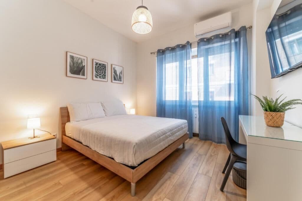 Een slaapkamer met een bed, nachtkastje, lamp, gordijnen en een potplant.