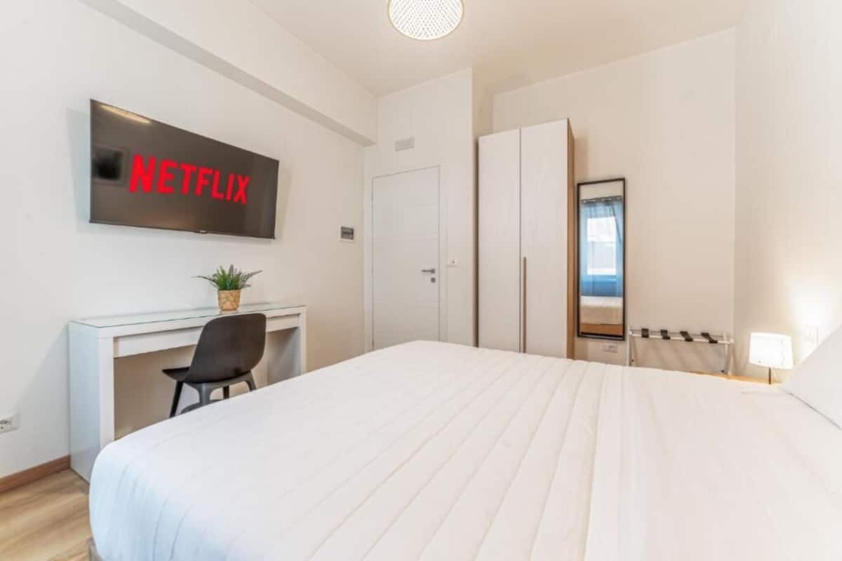 Ein Hotelzimmer mit einem Bett, einem Schreibtisch mit Stuhl, einem Kleiderschrank, einem Fernseher, der 'NETFLIX' anzeigt, und einem Fenster mit Vorhängen.