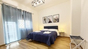 A bedroom with a bed, a bedside table with a lamp, a wall-mounted TV, and a drying rack.