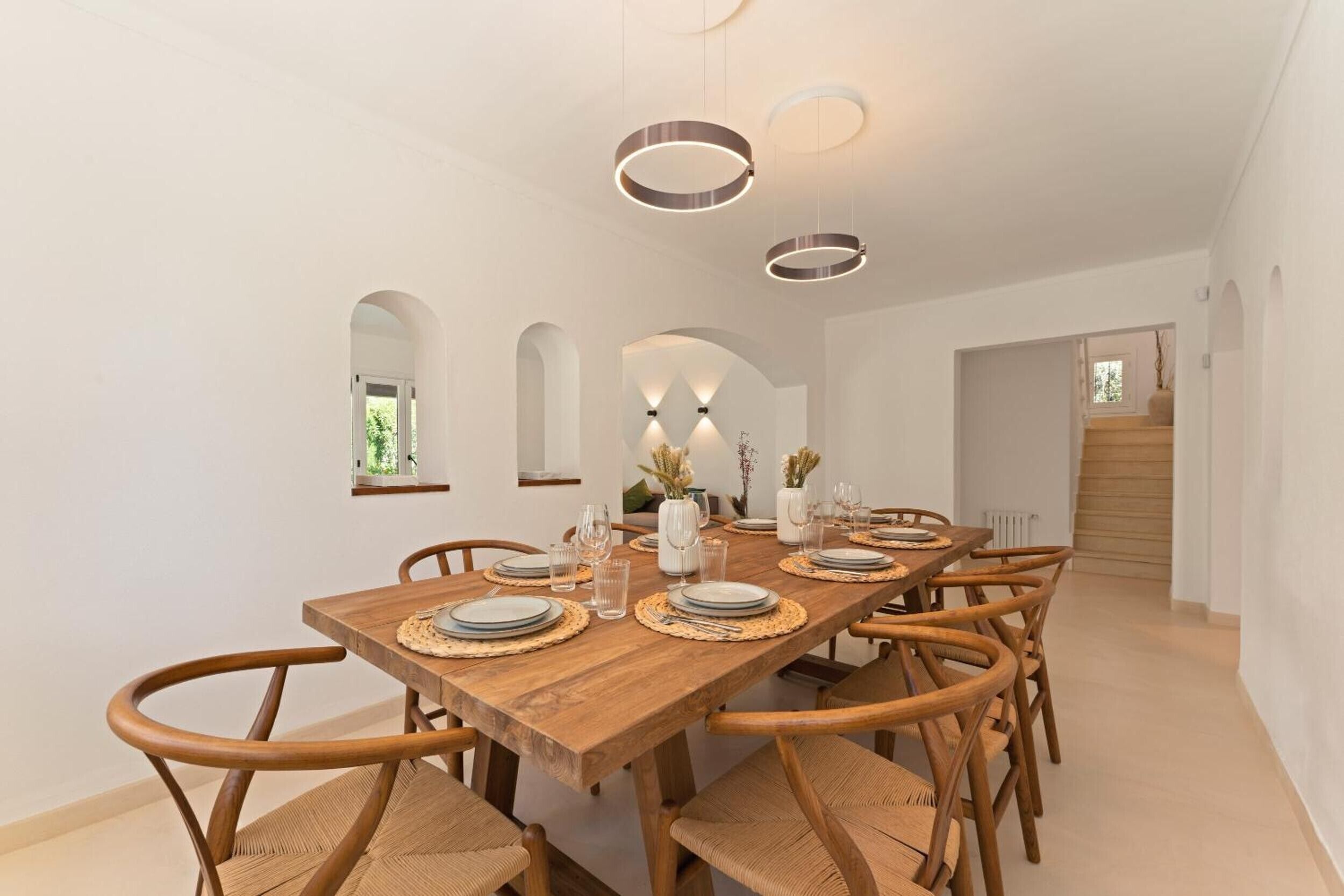 Uma sala de jantar com mesa de madeira posta para quatro pessoas, rodeada por cadeiras de vime, com iluminação pendente.