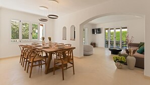 Ein geräumiger Essbereich mit einem Holztisch und Stühlen, einem an der Wand befestigten Fernseher und einem Balkon mit einer Couch und Pflanzen.