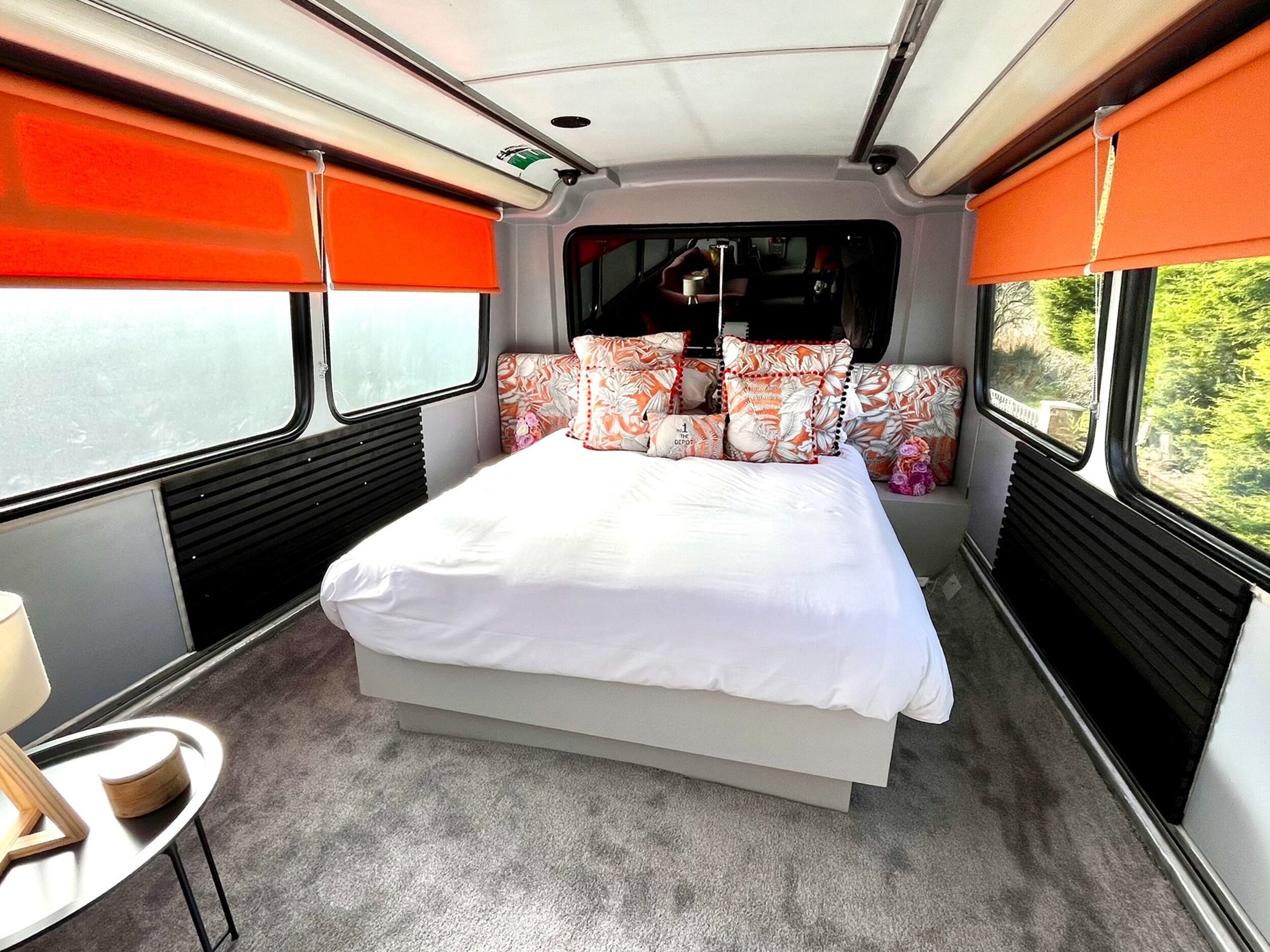 A bed with white bedding, orange and white patterned pillows, and a view of greenery through the window.