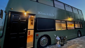 A double-decker bus with a 'Welcome to the Bus' sign, a door, windows, and a small table with a lamp and candle. - No 1 The Depot (Gateshead)