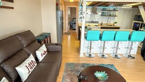 A living room with a brown sofa, a coffee table, and a kitchen area with bar stools. - The Little Penthouse is a charming two-level condo located in the gated Southwood Shores. This comfortable retreat offers access to resort style amenities including indoor and outdoor pools, a hot tub, playground, swimming area. (Lake Ozark)