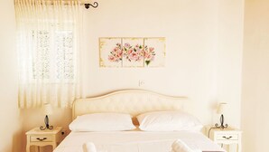 Un lit bien fait, avec du linge de lit blanc, deux tables de chevet avec des lampes, une œuvre d’art florale accrochée au mur et des rideaux en dentelle.