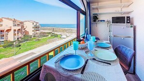 A balcony with a table set for dining, a view of the sea, and a kitchen area with a refrigerator and microwave. - Palm Beach by Interhome (Le Barcarès)
