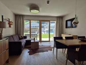 Ett modernt vardagsrum med en soffa, ett matbord med stolar, en TV som är monterad på väggen och en skjutdörr i glas som leder ut till en trädgård.