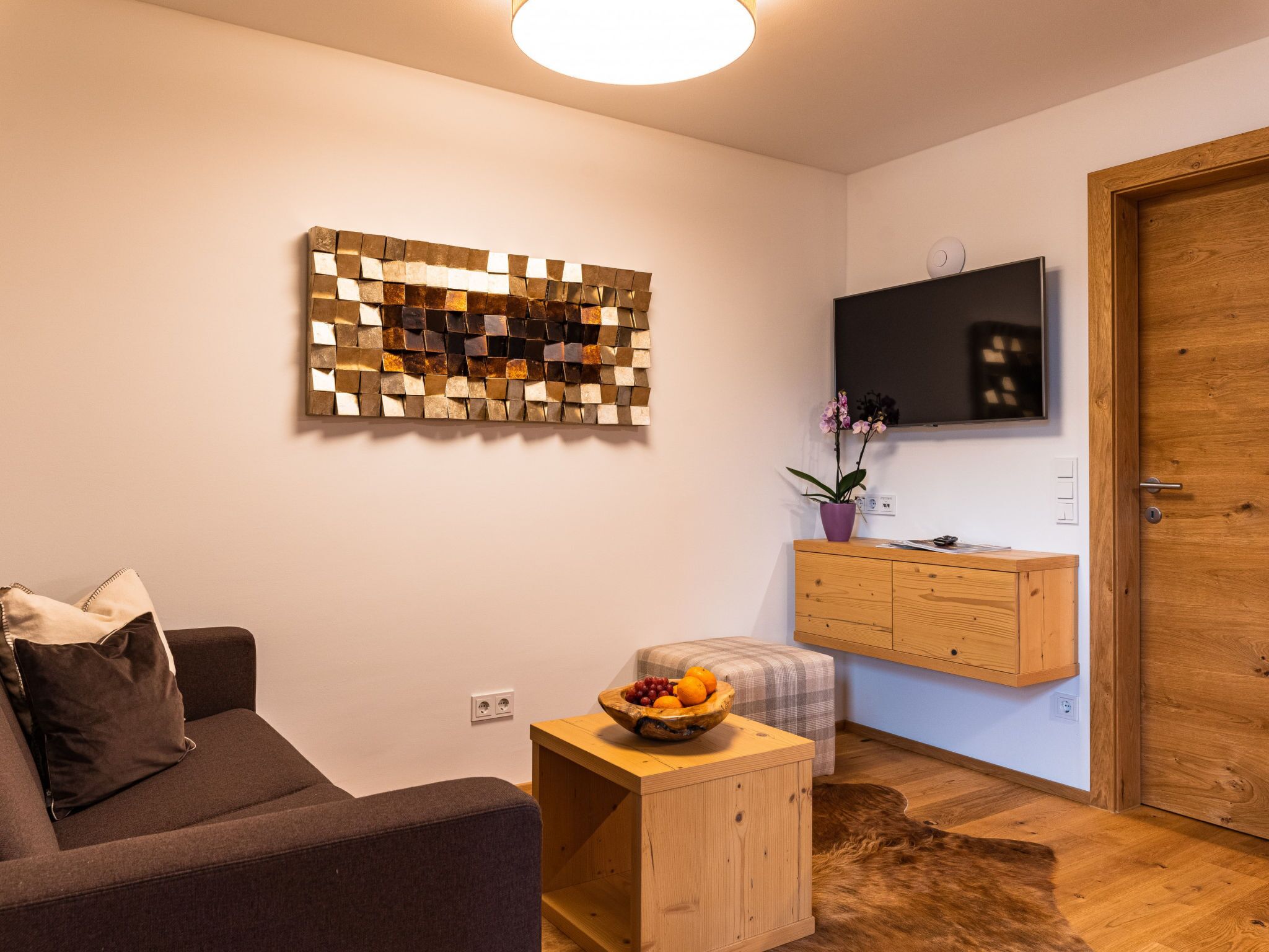 Un salon moderne comprenant un canapé, une table d’appoint en bois avec des fruits, un élément décoratif mural, un téléviseur fixé au-dessus d’un meuble en bois et une porte en bois.