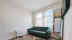 A room with a herringbone-patterned wooden floor, a green sofa, a small white side table, and a large mirror reflecting a door and window.