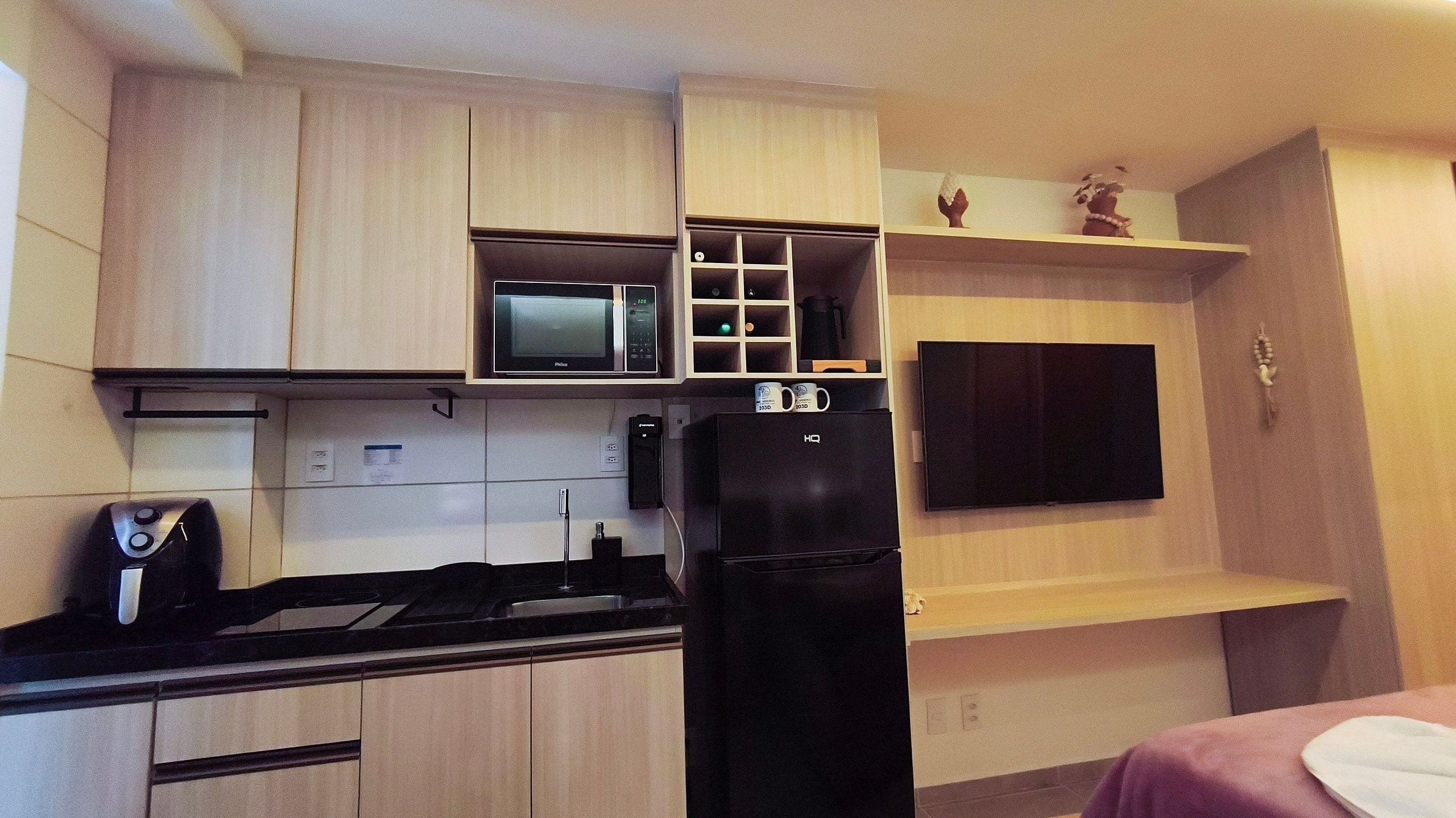 A compact kitchen with a microwave, a black refrigerator, and a flat-screen TV mounted on the wall.