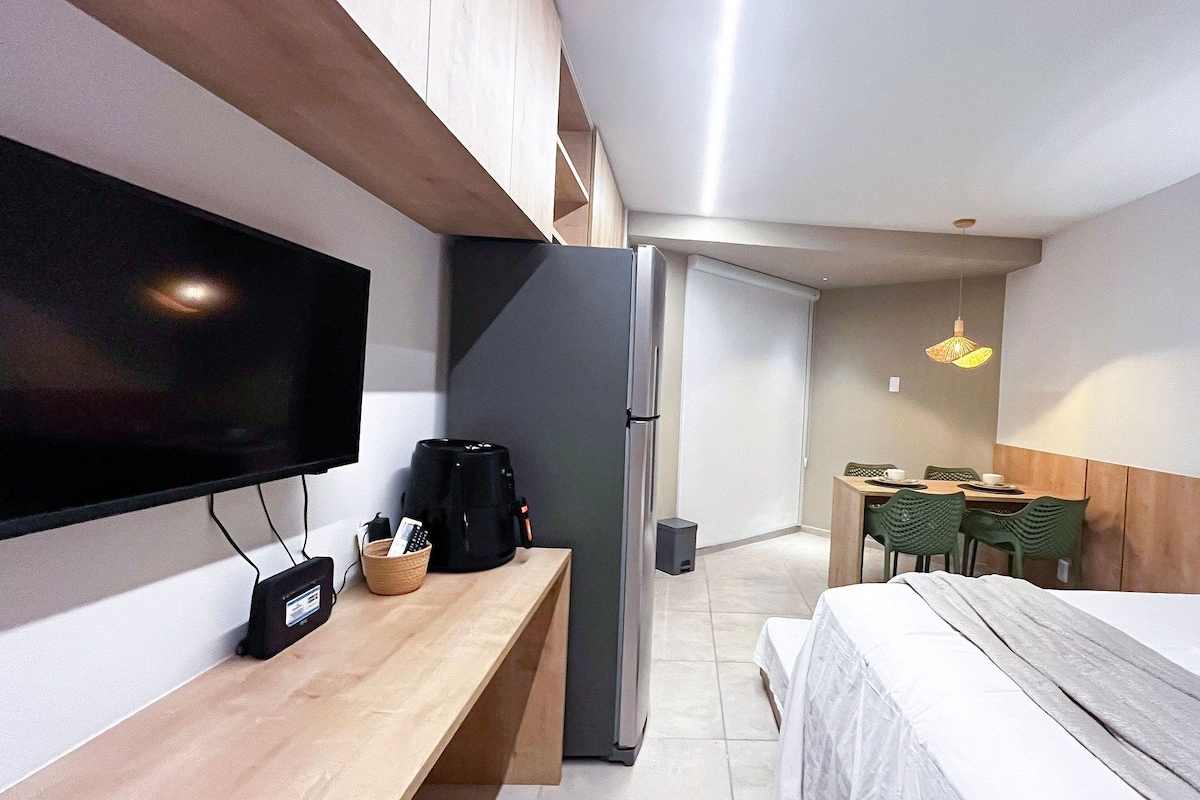 Quarto de hotel pequeno e funcional, com uma cama, escrivaninha, televisão, cafeteira e área de refeições com cadeiras.