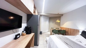 Quarto de hotel pequeno e funcional, com uma cama, escrivaninha, televisão, cafeteira e área de refeições com cadeiras.
