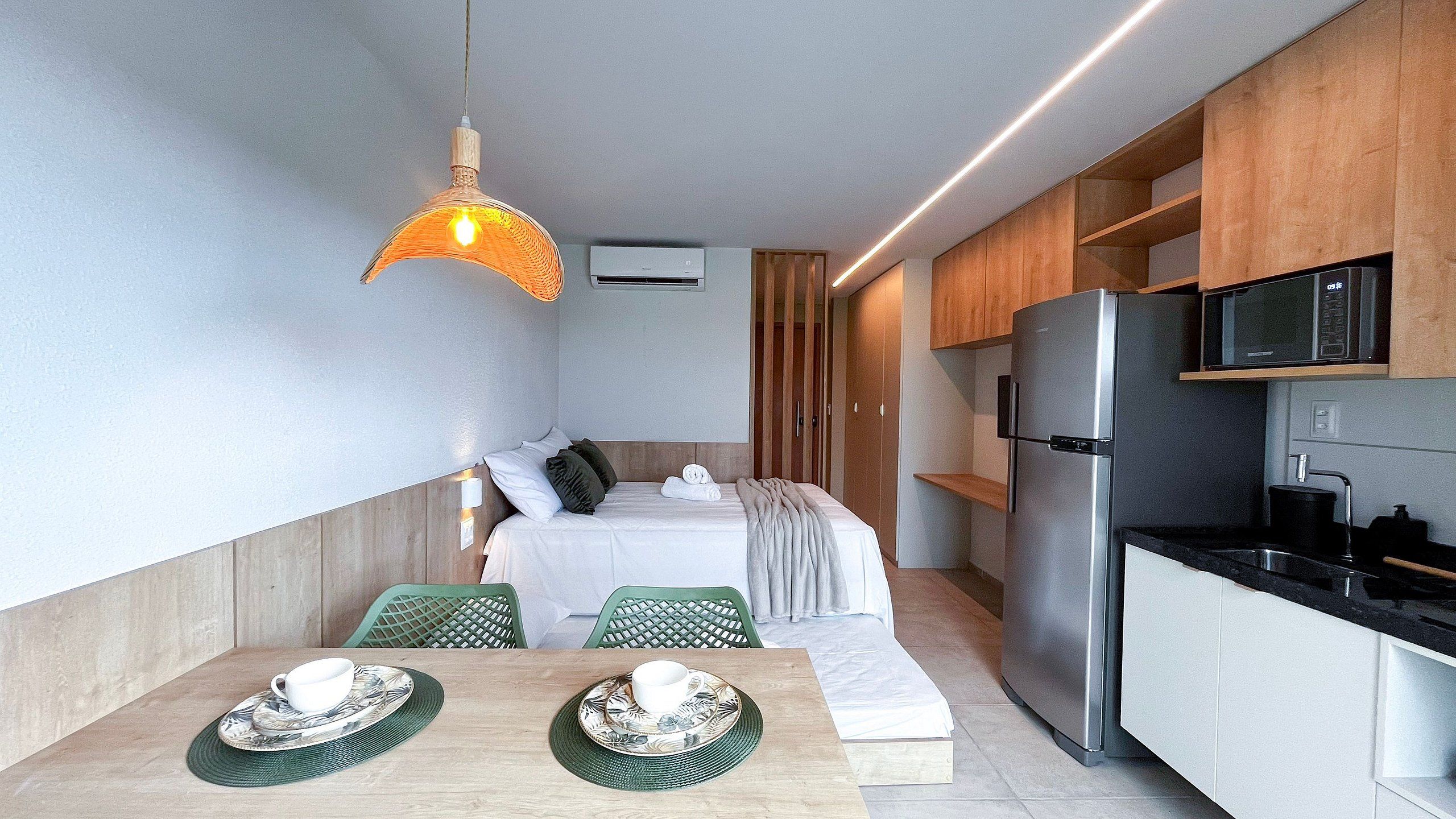 A modern hotel room with a bed, a dining table with chairs, a kitchen area with a refrigerator and microwave, and a hanging light fixture.