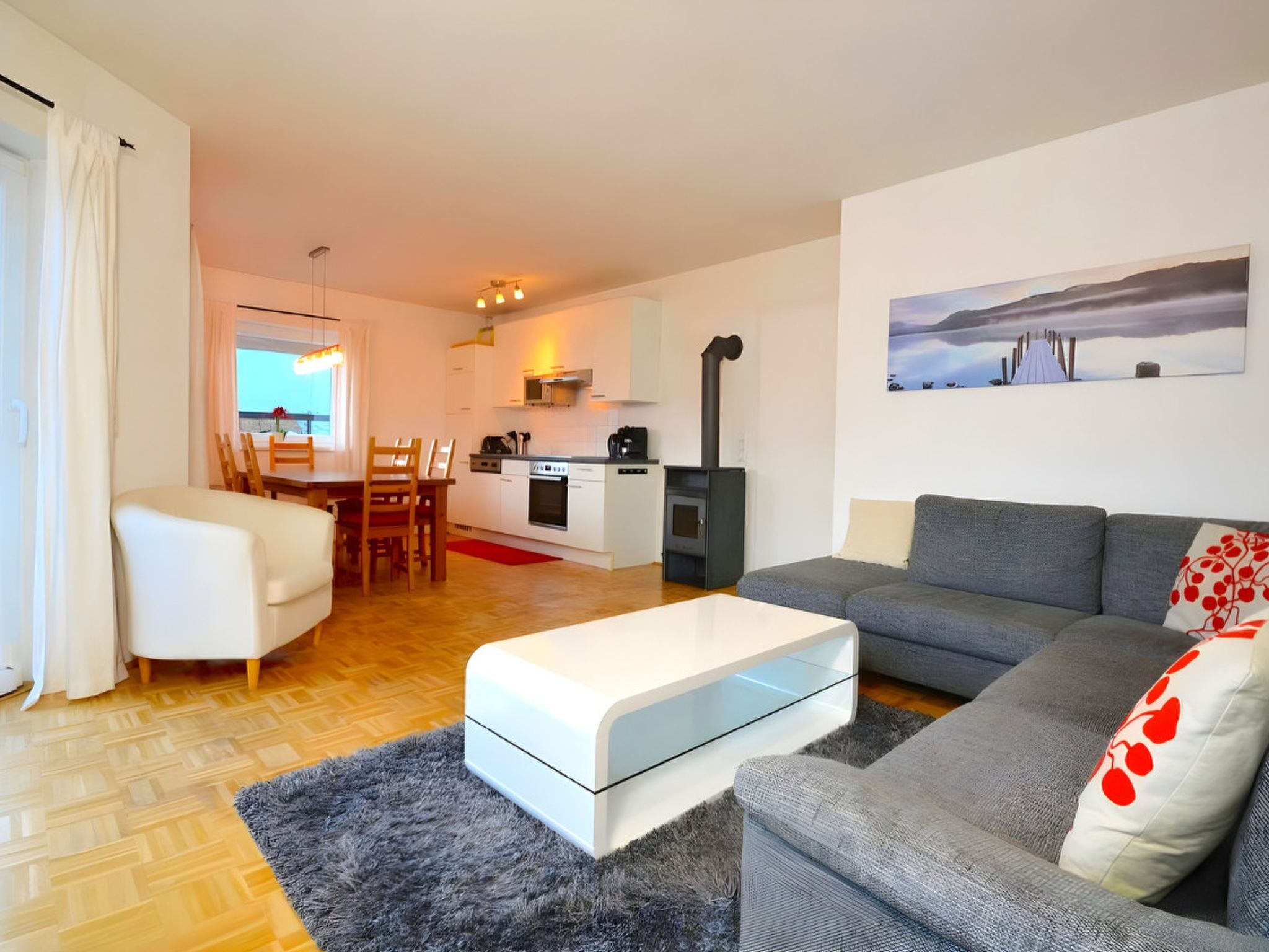 A modern living room with a sofa, a dining area with chairs, a kitchen with a stove, and a large window.