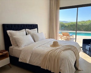 Un dormitorio con una cama grande, una mesita de noche, una silla y vistas a una piscina.