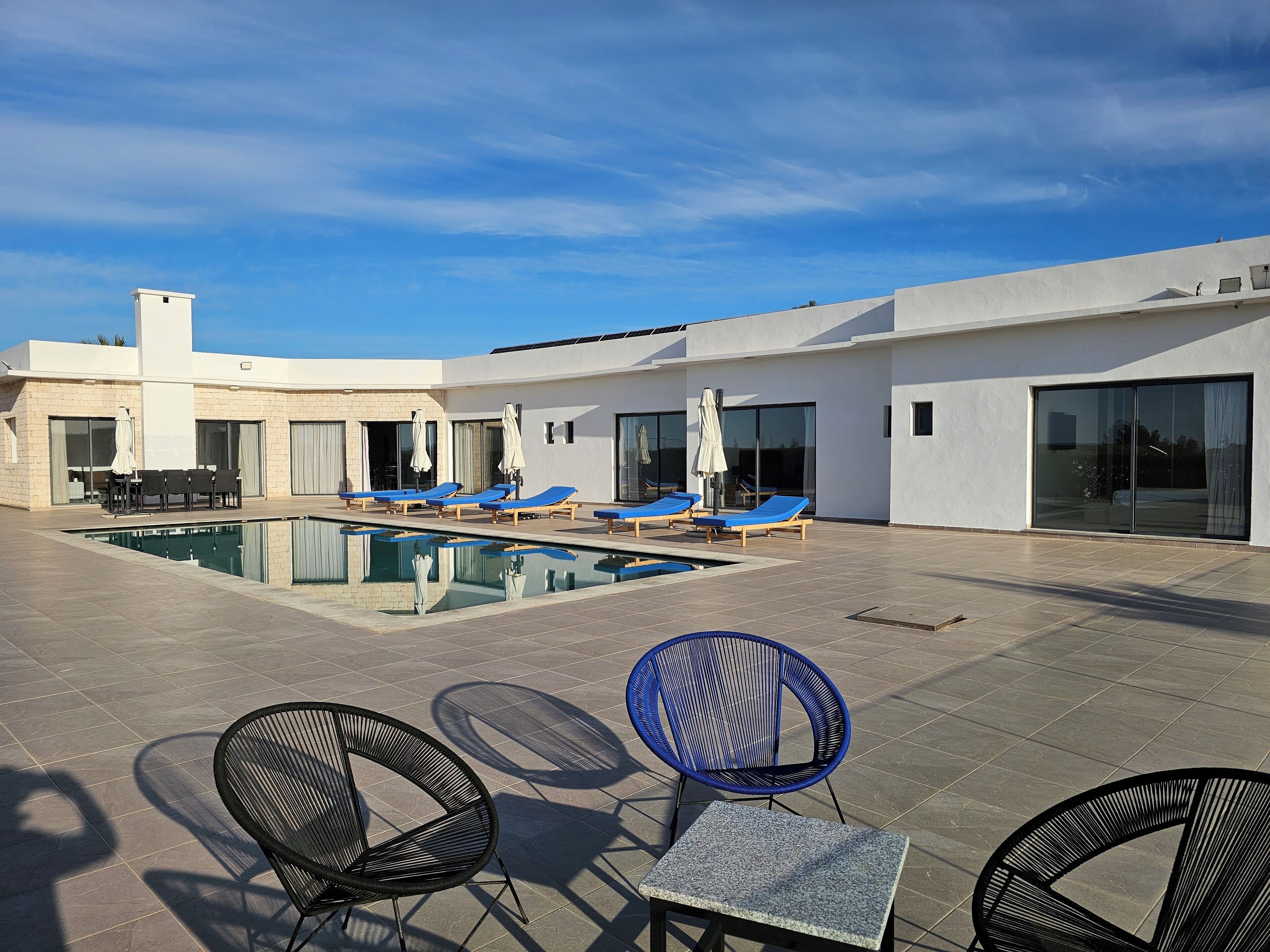 A modern outdoor pool area with lounge chairs and a table, surrounded by a building with large windows and a flat roof.