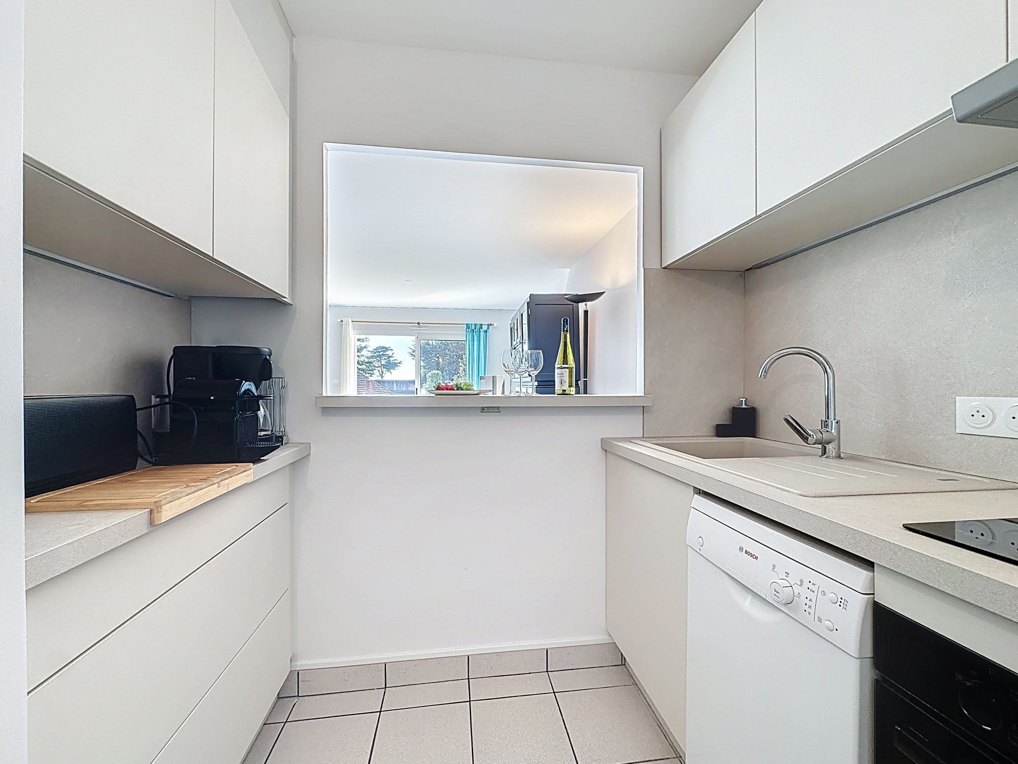A compact kitchen with white cabinets, a Bosch dishwasher, a coffee machine, and a window with a view of the outside.