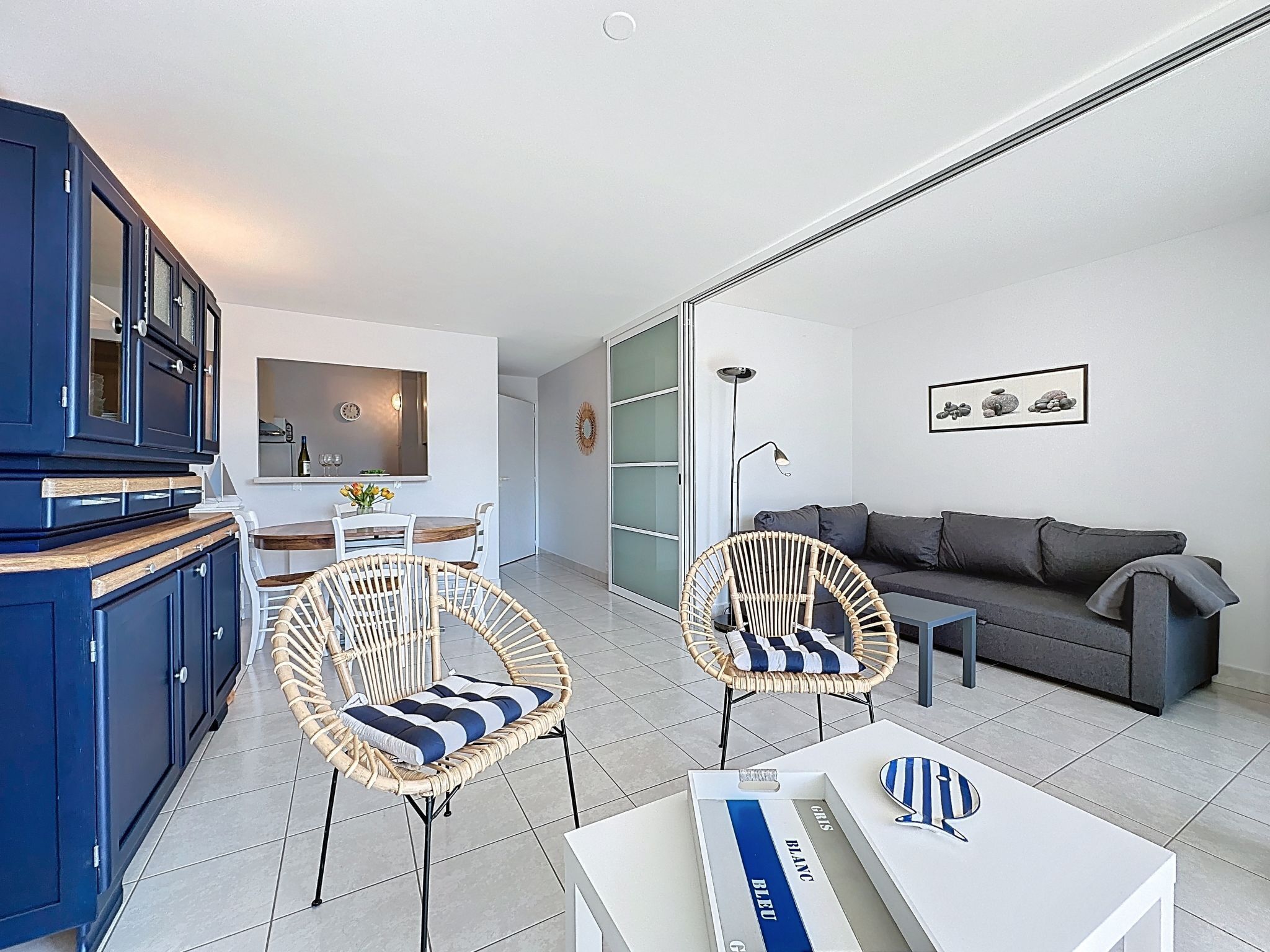 Un salon moderne comprenant un canapé, deux fauteuils en osier, une table  à manger avec des chaises, un coin cuisine avec des placards et un grand miroir.