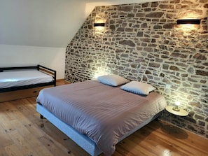 A bedroom with a stone wall, a bed with pillows, a bedside table with a lamp, and a wooden floor. - Holiday Home “Petit Loft, Maison Bord De Mer” with Private Garden and Wi-Fi (Pirou)