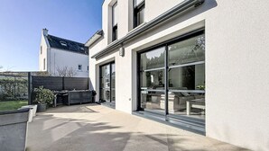 Ein modernes Haus mit einer großen Glasdoor, einer Terrasse mit Gartenmöbeln und einem sichtbaren Gartenbereich.