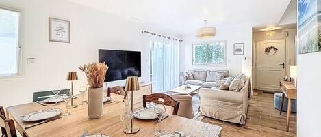 A modern living room with a dining table set for four, a sofa, a TV, and a door with a wreath.