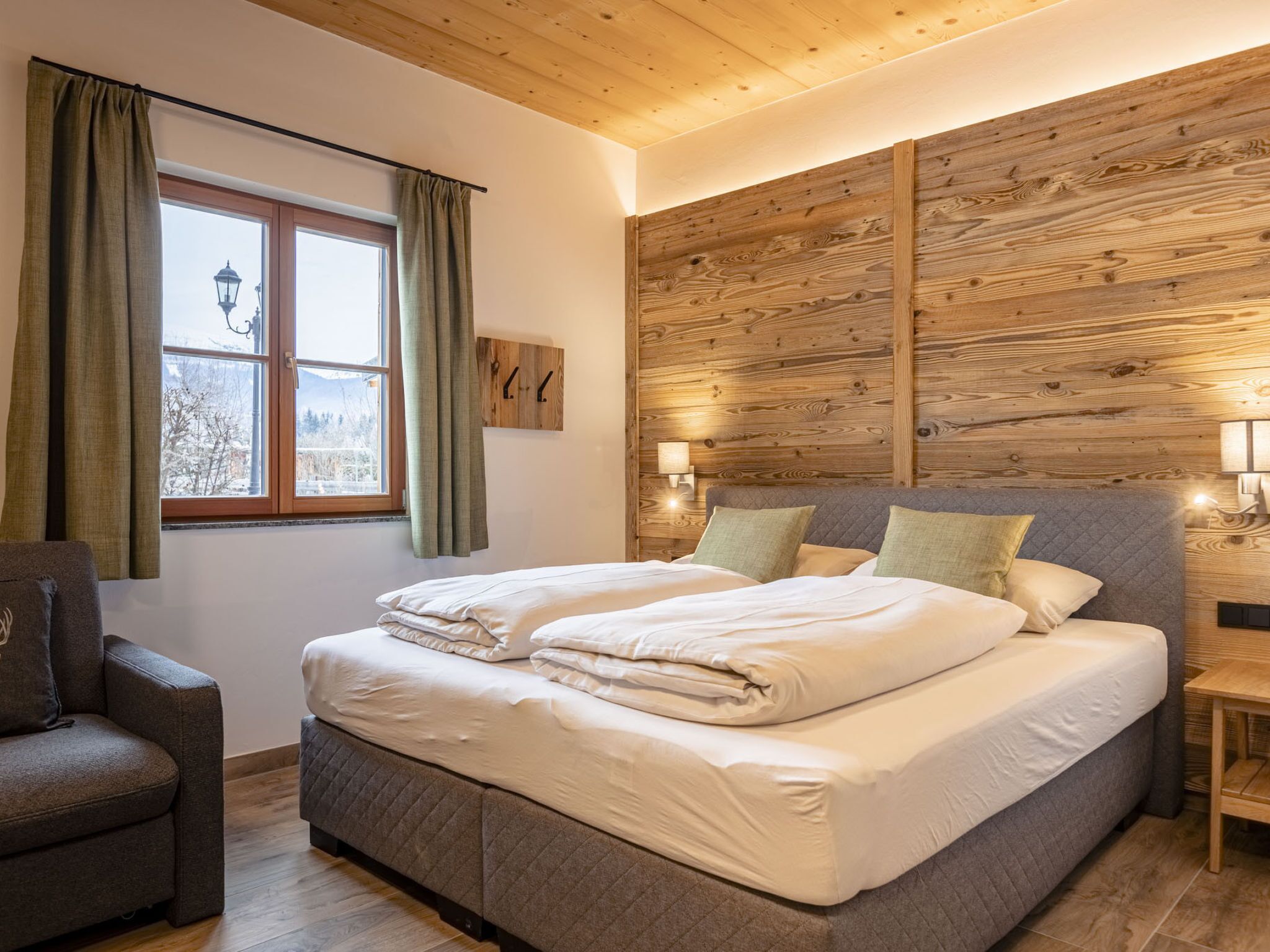 A bedroom with a large bed, a window with curtains, a wall-mounted lamp, and a wooden headboard.