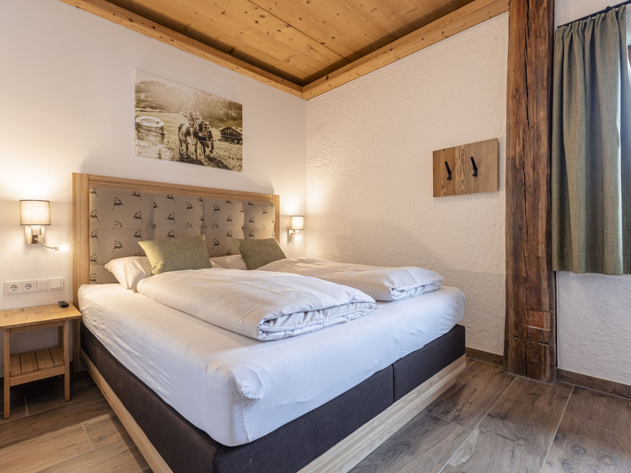A double bed with a wooden headboard, two bedside tables, a wall-mounted lamp, and a painting above the bed.