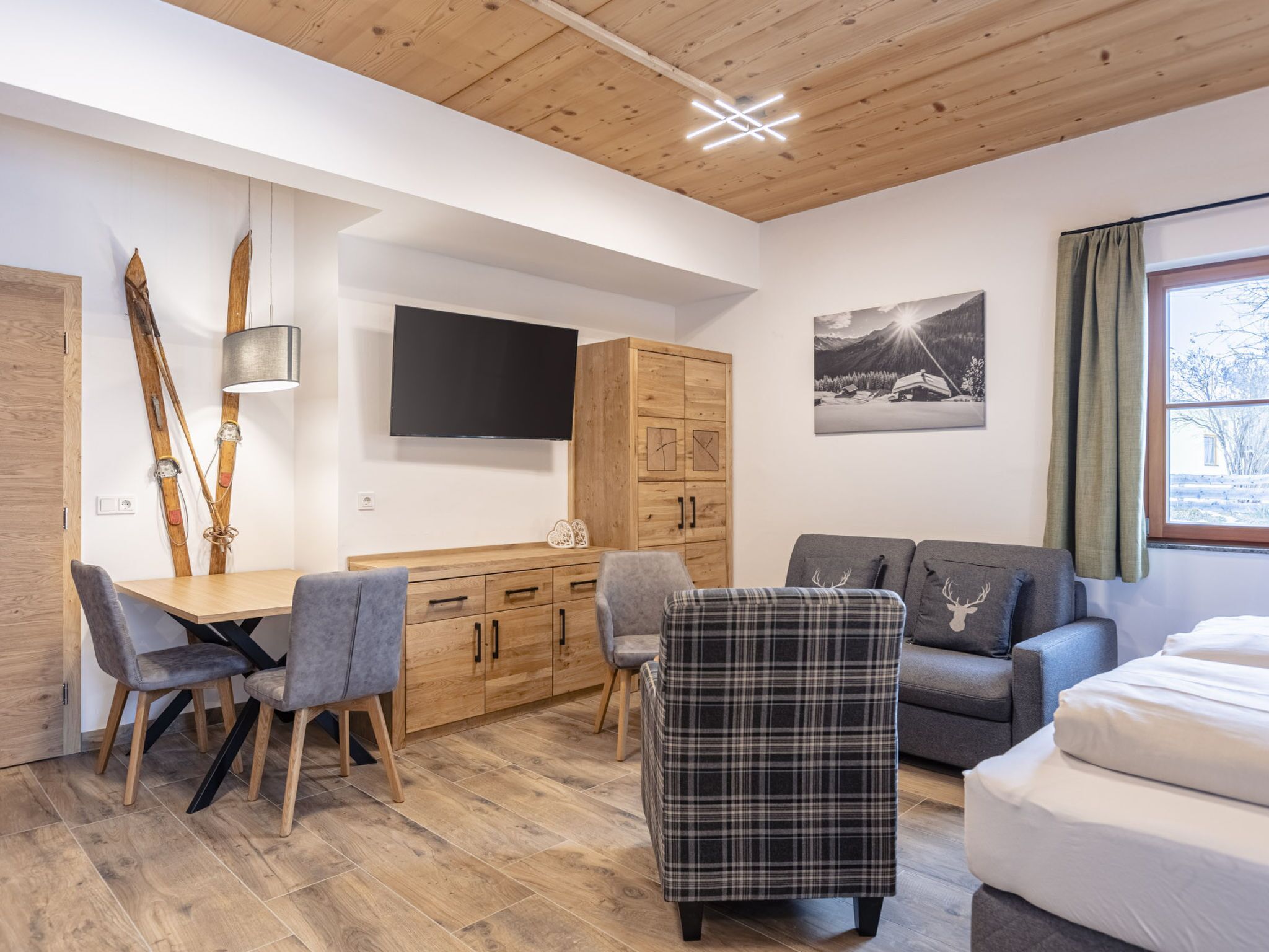 Una stanza con soffitto in legno, una TV montata a parete, un armadietto in legno, un divano, una poltrona a quadri e un tavolo da pranzo con sedie.