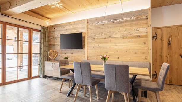 Ein modernes Interieur mit Holzboden, einem Wand-Fernseher, einem Essbereich mit grau gepolsterten Stühlen und einem Holztisch sowie einer Glastür zum Außenbereich.