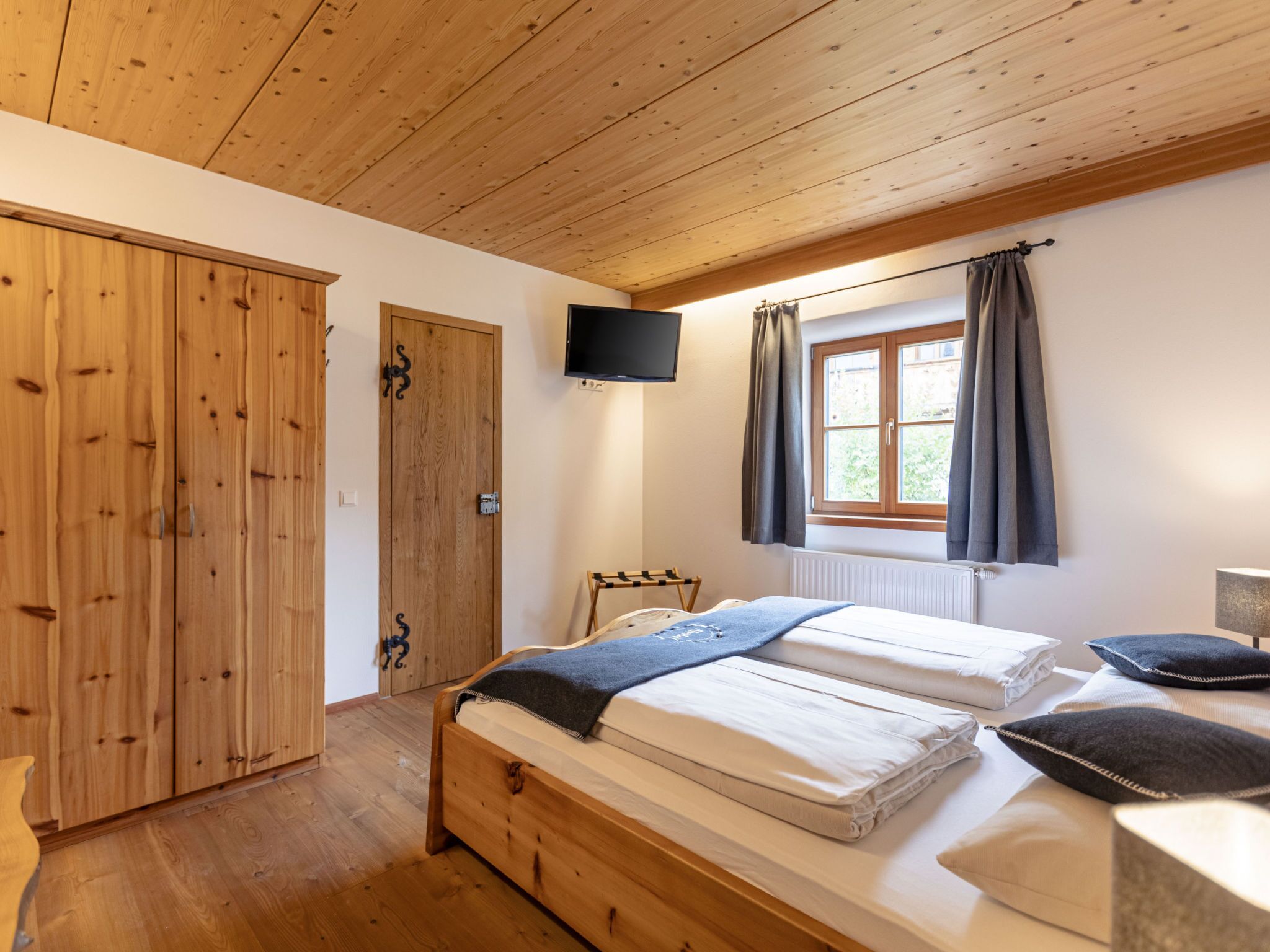 A bedroom with a wooden ceiling, a bed with white bedding, a TV mounted above a window with curtains, and a wardrobe with a mirror.