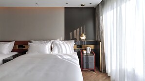 A hotel room with a large bed, a bedside table with a lamp, a small table with a coffee pot, and a view of the outdoors through sheer curtains. - Life Style  (Douala)