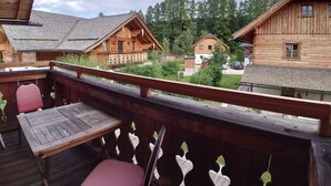 Um deck de madeira com mesa e cadeiras, com vista para uma fileira de cabanas de toras tradicionais com telhados inclinados e um cenário de floresta.