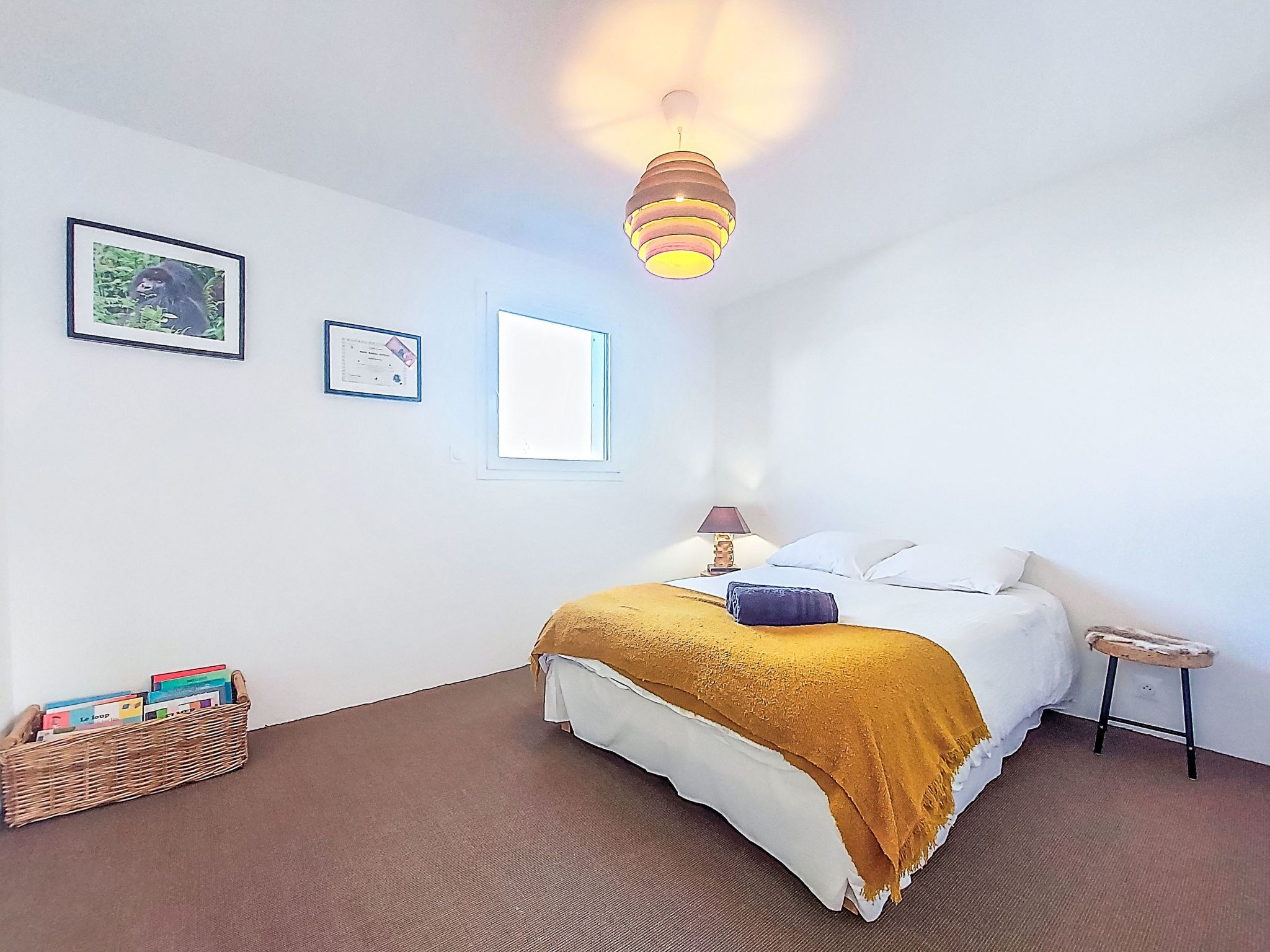 Une chambre à coucher comprenant un lit, une table de chevet, une lampe, un luminaire suspendu, une fenêtre et un certificat accroché au mur.
