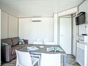 Ein Zimmer mit grauem Sofa, weißen Stühlen, einem Tisch mit Zeitschriften, einem an der Wand montierten Fernseher und einer Tür.