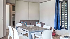 Ein modernes Zimmer mit Esstisch, weißen Stühlen, einer grauen Couch mit bunten Kissen, einem blauen Vorhang und einem Fenster mit Jalousien.