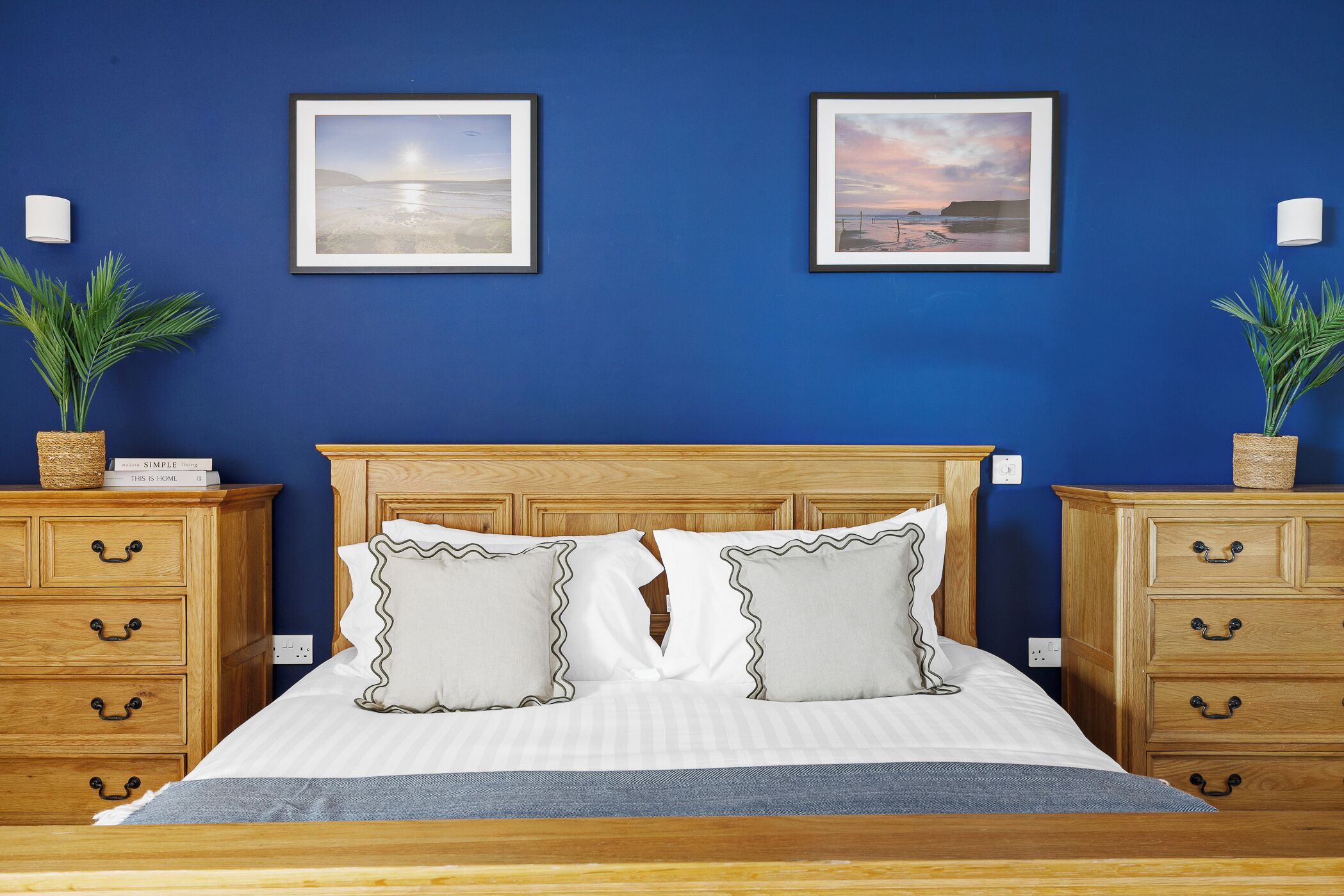 A bedroom with a wooden headboard, two bedside tables with lamps, a bed with white bedding, two framed pictures on the wall, and two potted plants.