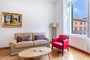 Ein Zimmer mit einer Couch, einem roten Sessel, einem Tisch mit Tassen, einer Lampe und einem Fenster mit Blick auf Gebäude.