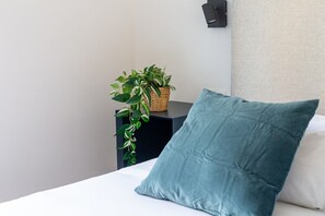 Una cama con un cojín color turquesa, una mesita de noche negra con una cesta tejida que contiene una planta, y una lámpara de pared.