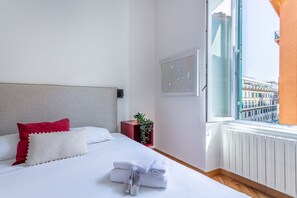 Ein Hotelzimmer mit Bett, Kissen, einem roten Nachttisch, einem gerahmten Bild und einem Fenster mit Blick auf die Stadt.