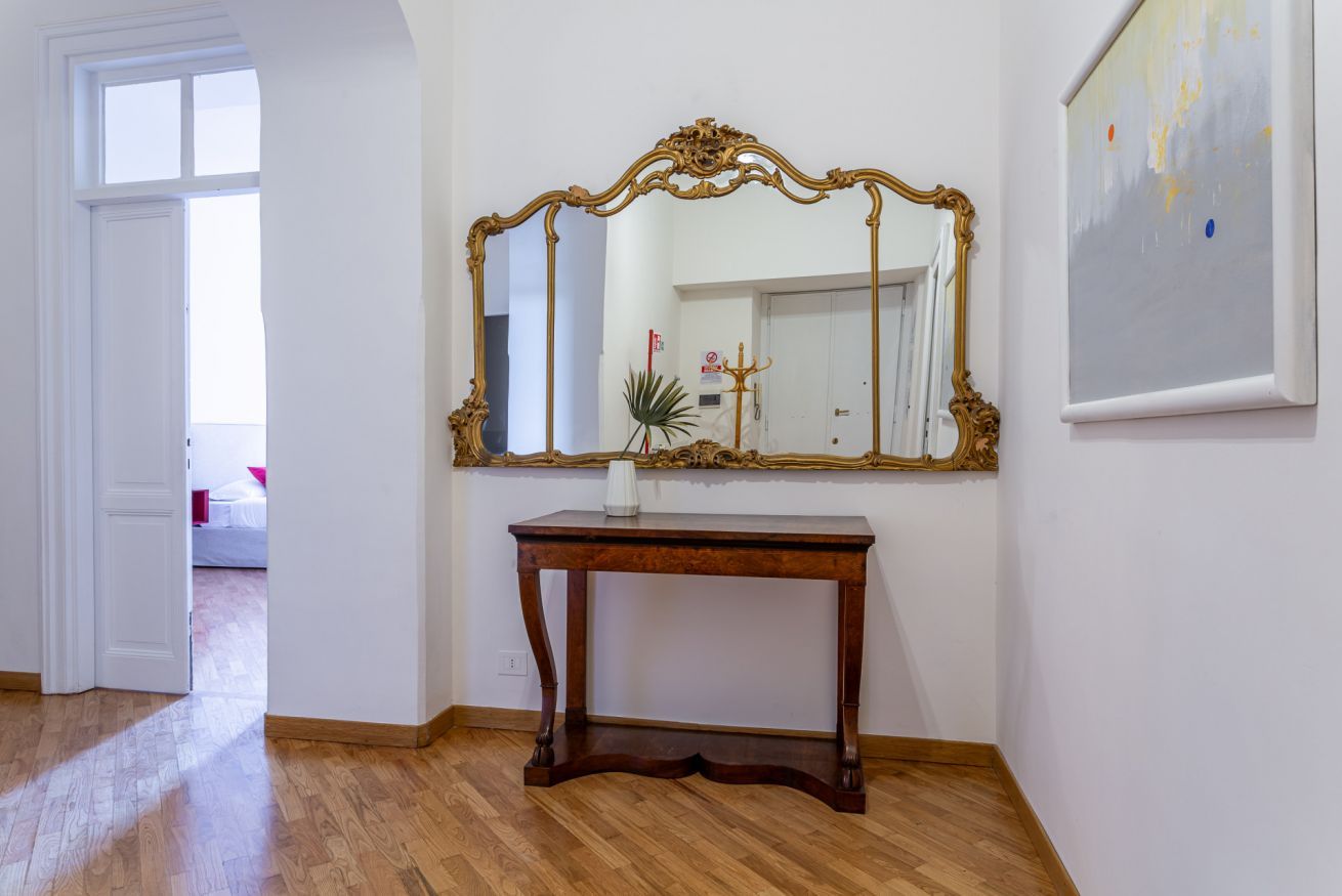 A room with a wooden floor, a large ornate mirror on the wall, a small table with a vase, and a painting.