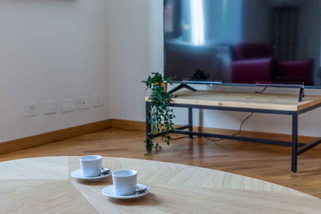 A room with a wooden floor, a table with two white cups, a plant, and a large TV on a stand.