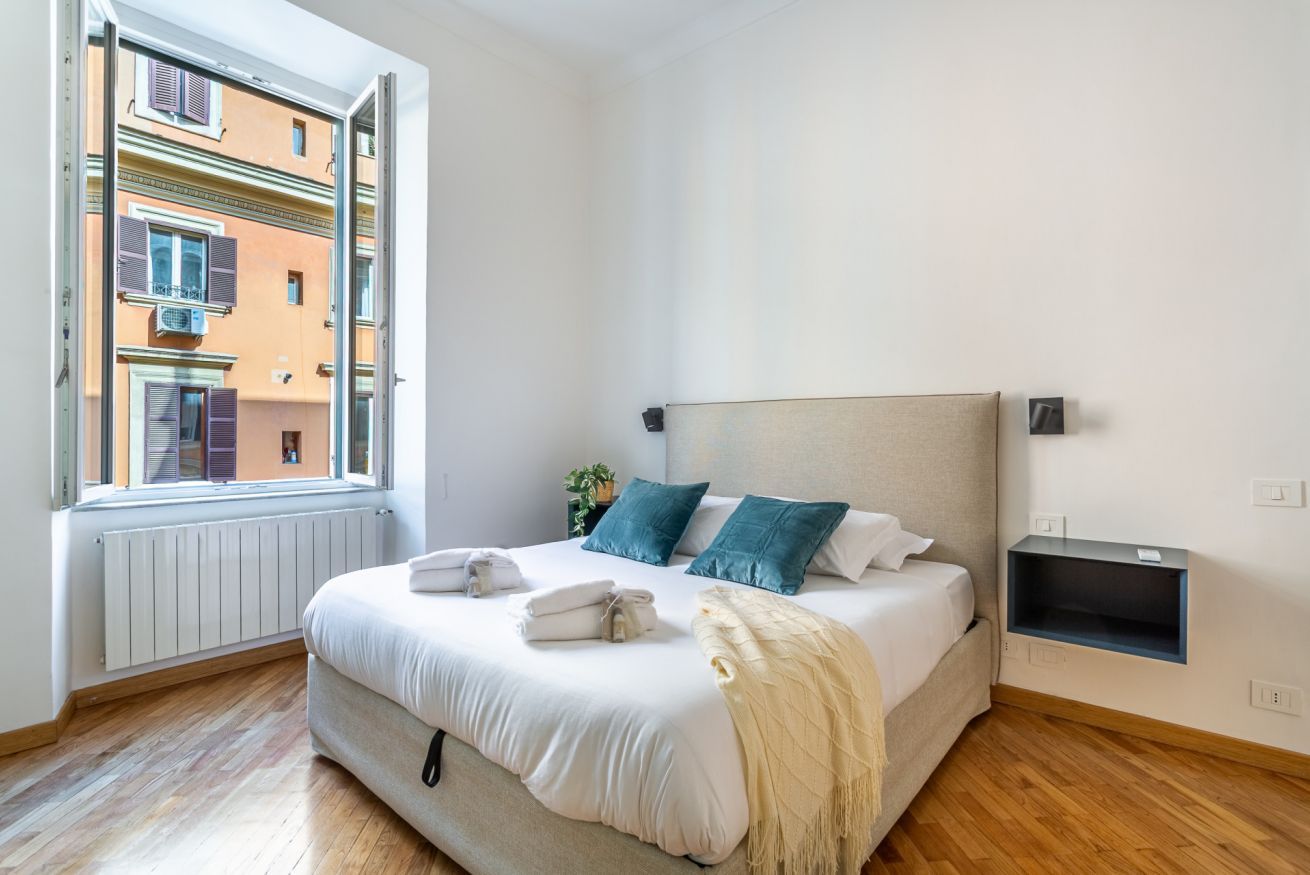 A bedroom with a bed, a window, a radiator, and a small shelf.