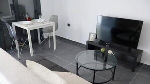 A modern living room with a sofa, a glass coffee table, a TV, a dining table with chairs, and a small side table.
