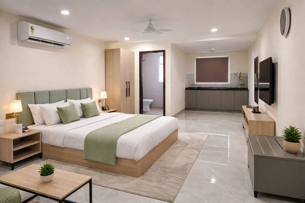 A modern hotel room with a bed, bedside tables, a desk, a TV, and a kitchenette.
