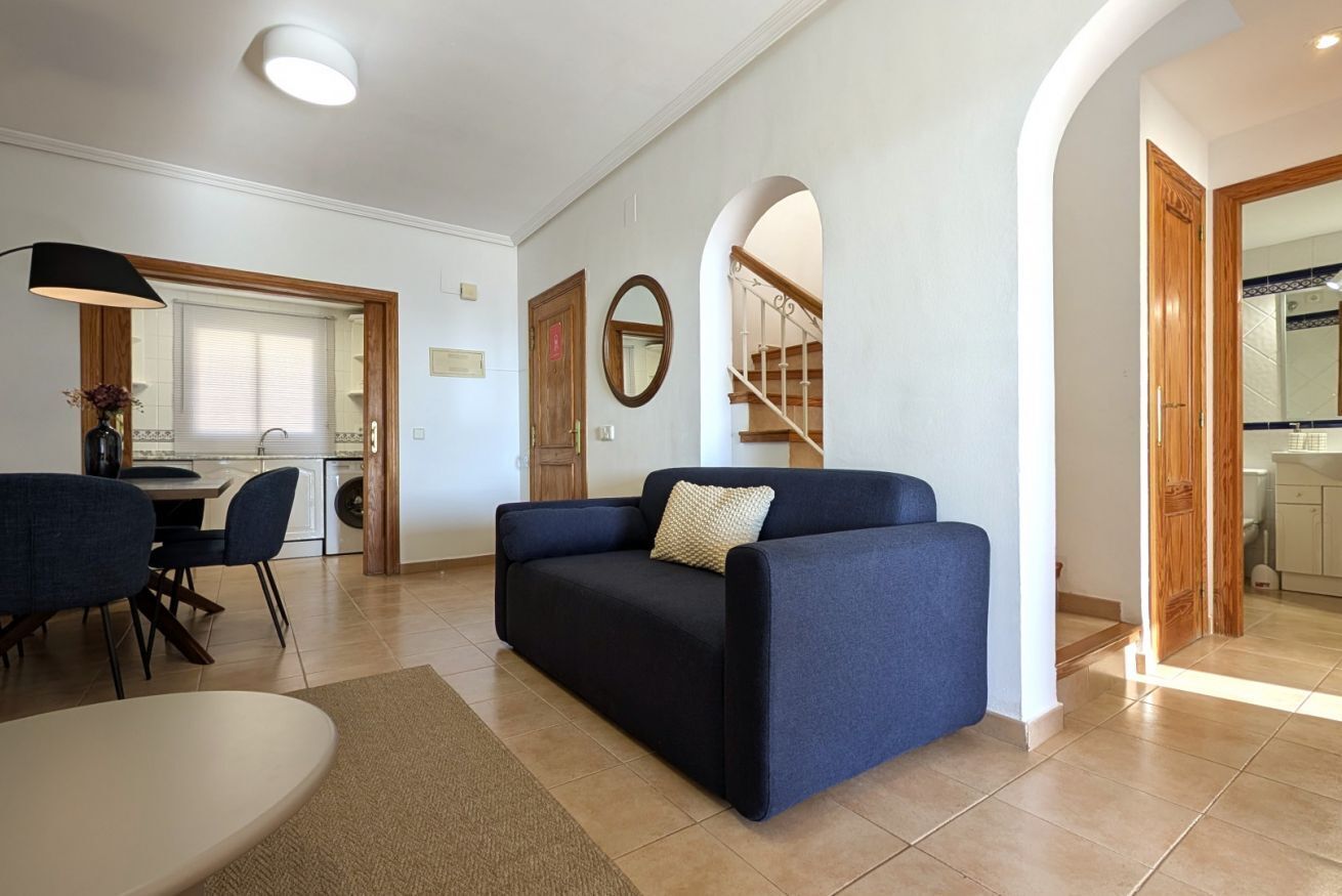 A living room with a sofa, a dining table, and chairs. There is a staircase leading to an upper floor.