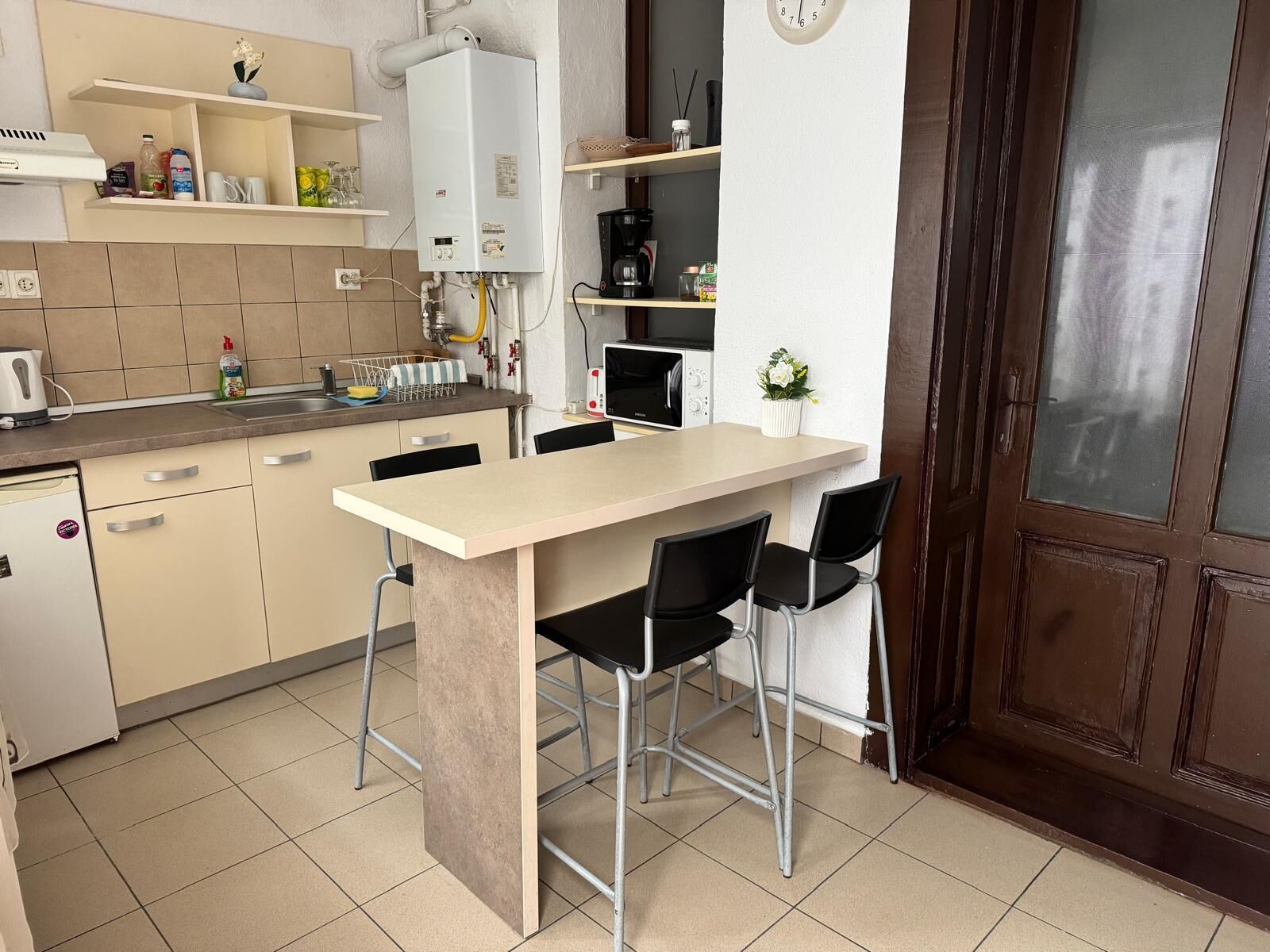 A compact kitchen with a dining area, a white refrigerator, a coffee maker, and a clock on the wall.